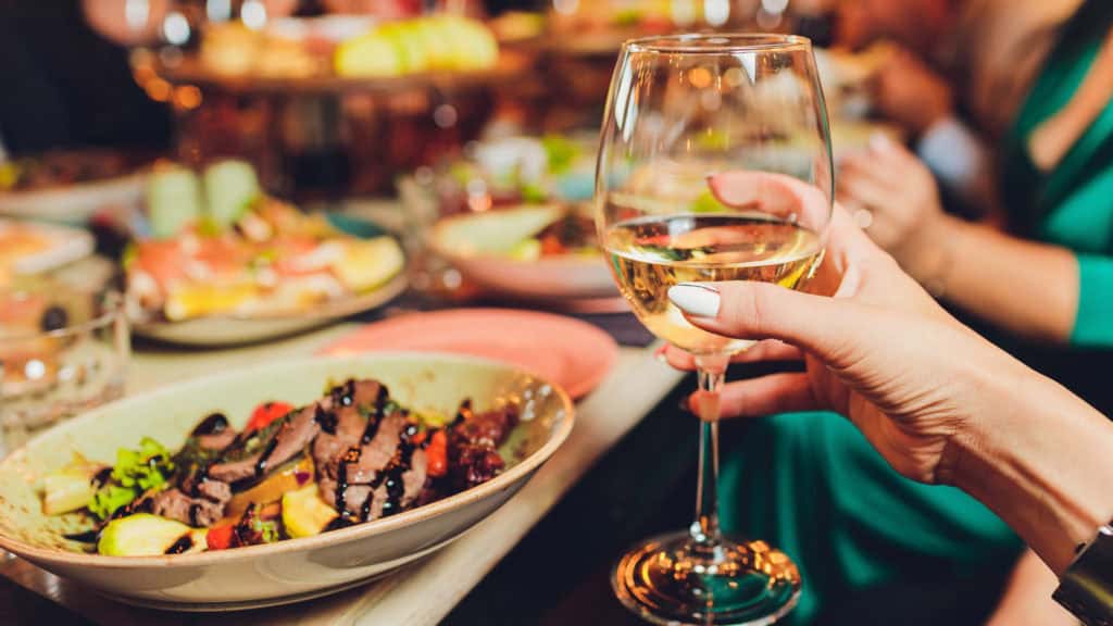 Hand mit weiss lackierten Fingernägeln hält ein Weinglas mit Chardonnay vor einem Teller mit Salat und Fleischstreifen, im Hintergrund unscharfe Personen und weitere Teller