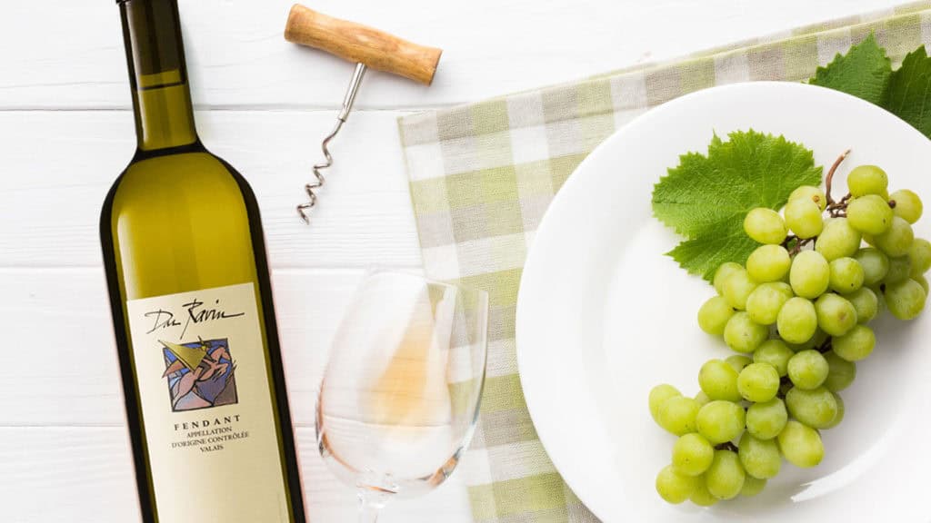 Flasche Chasselas-Weisswein mit Korkenzieher, Weinglas und Teller mit grünem Traubenbund und Weinblatt auf kariertem Tuch