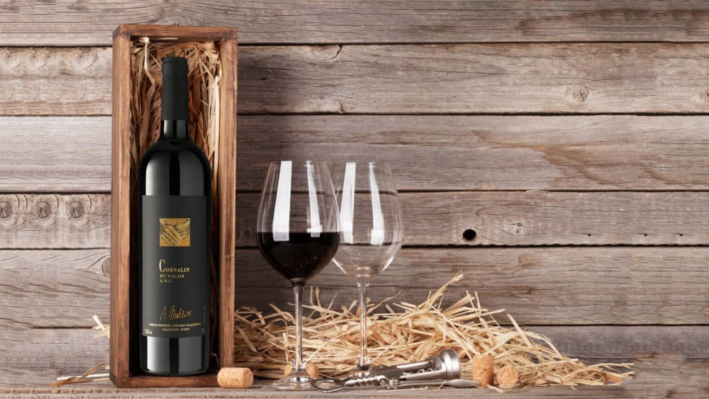 Flasche Cornalin Adrian Mathier in einer Holzbox neben zwei Weingläsern, Korken und Weinverschluss auf Holzuntergrund