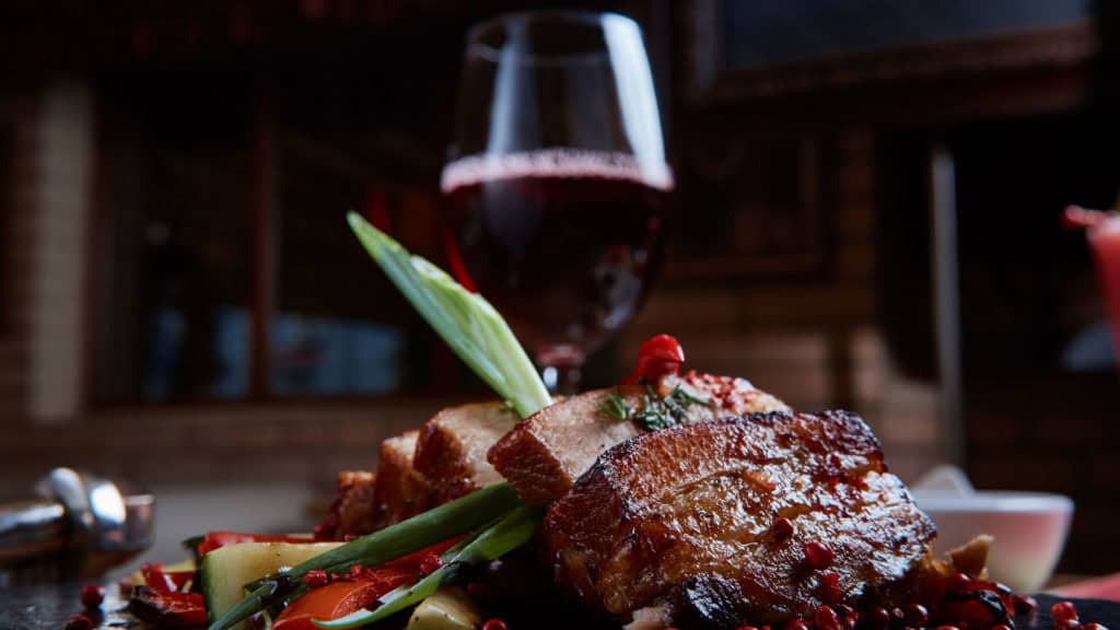 Wildschweingericht mit knuspriger Kruste, garniert mit Frühlingszwiebeln und rotem Chili, im Hintergrund ein Glas Cornalin