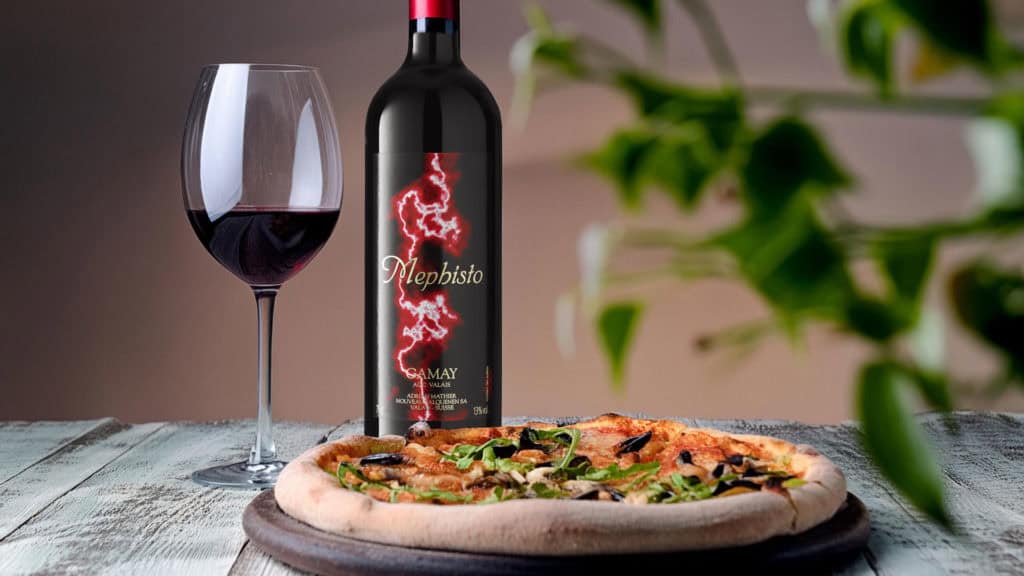 Flasche Gamay mit Etikett 'Mephisto Gamay', daneben ein Glas Rotwein und eine Pizza mit grünem Belag auf einem Holztisch