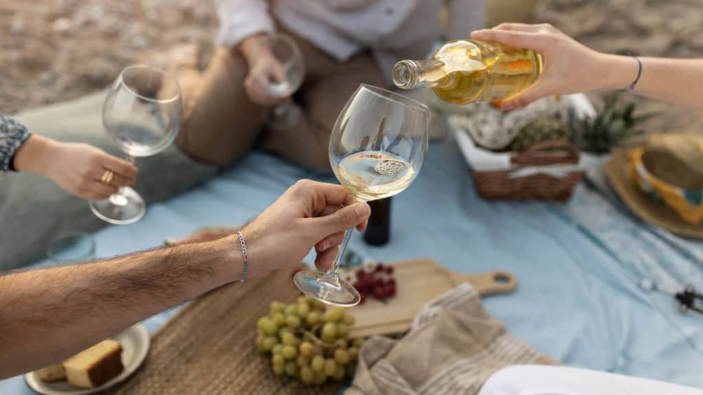 Person giesst Humagne Blanc in ein Weinglas bei einem Picknick im Freien mit Trauben und Brot auf einer blauen Decke
