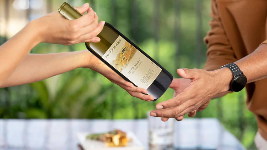Zwei Hände übergeben eine Weinflasche mit Etikett 'Les Pyramides Petite Arvine' vor unscharfem Hintergrund mit Glas und Teller