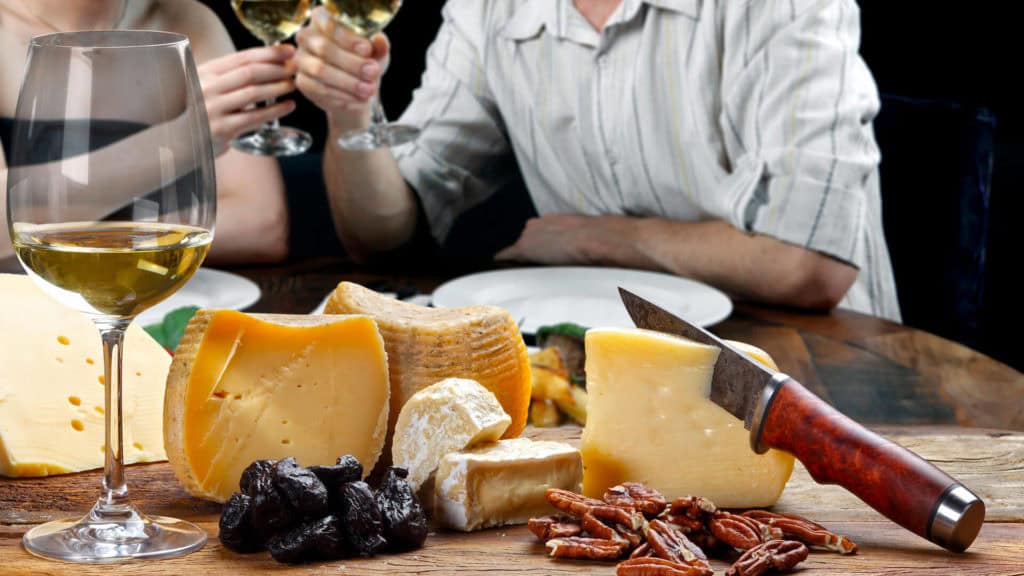 Käseplatte mit verschiedenen Käsesorten, Trockenfrüchten und Nüssen auf einem Holztisch, daneben ein Glas Weisswein Petite Arvine und zwei Personen, die sich mit Weingläsern zuprosten