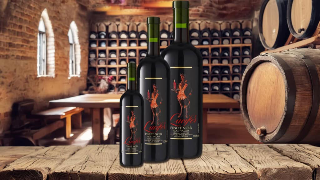 Drei unterschiedlich grosse Flaschen Pinot Noir mit schwarzem Etikett und roter Illustration stehen auf einem Holztisch in einem Weinkeller mit Backsteinwänden und Weinfässern.