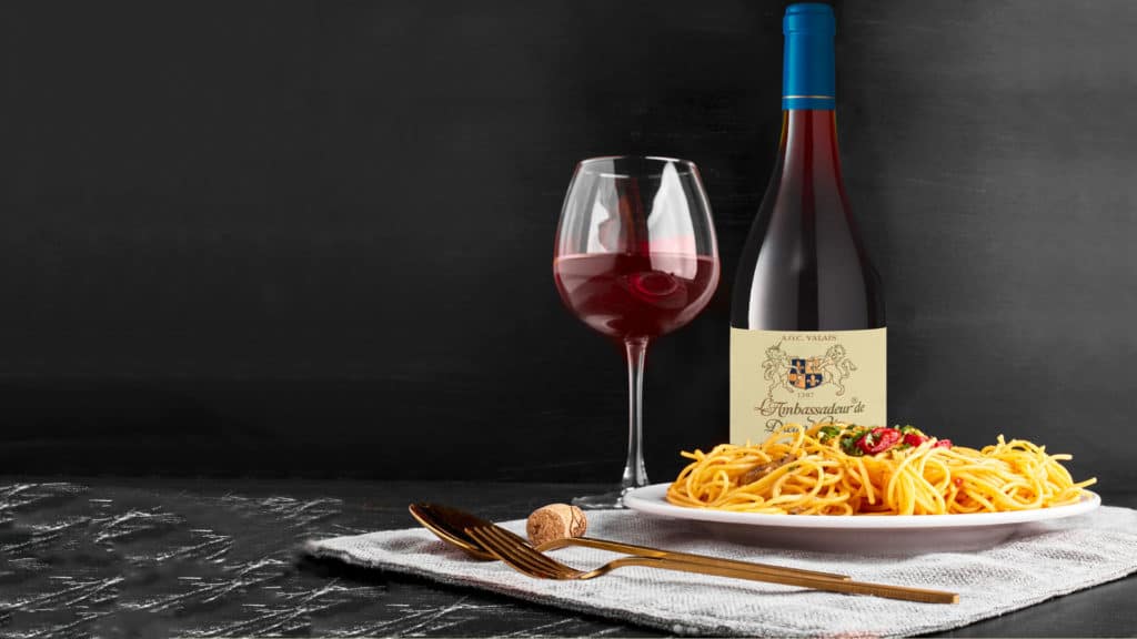 Weinglas mit Rotwein, Flasche Pinot Noir und Teller mit Spaghetti auf grauem Tuch vor schwarzem Hintergrund