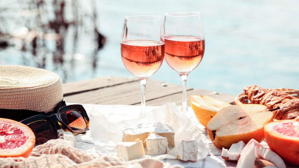 Zwei Gläser Roséwein auf einem Tisch mit Obst, Käse und Sonnenbrille für einen entspannten Tag am Wasser.
