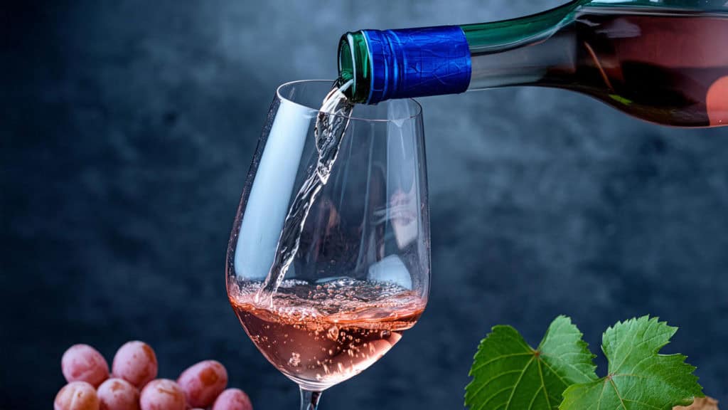 Ein Glas mit Roséwein wird aus einer Flasche eingeschenkt, umgeben von Trauben und Weinblättern.