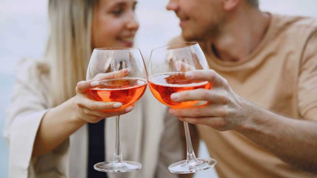 Paar prostet mit Roséwein in grossen Gläsern, lächelnd am Strand bei Sonnenuntergang.