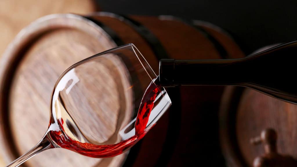 Rotwein wird aus einer Flasche in ein schräg gehaltenes Weinglas vor einem Holzfass eingeschenkt