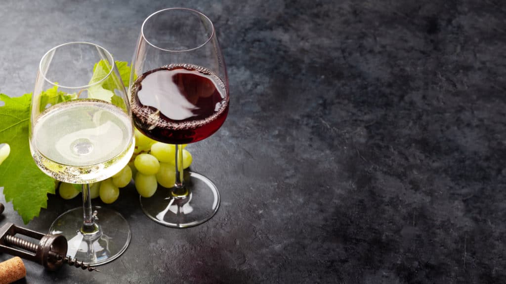 Zwei Weingläser mit Weiss- und Rotwein neben grünen Weintrauben und Weinblatt auf dunklem Untergrund