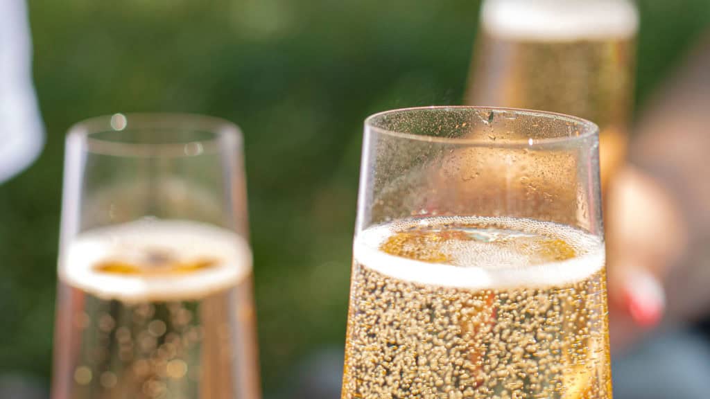 Sektgläser mit sprudelndem Schaumwein und Blasen, im Hintergrund grüne Wiese.