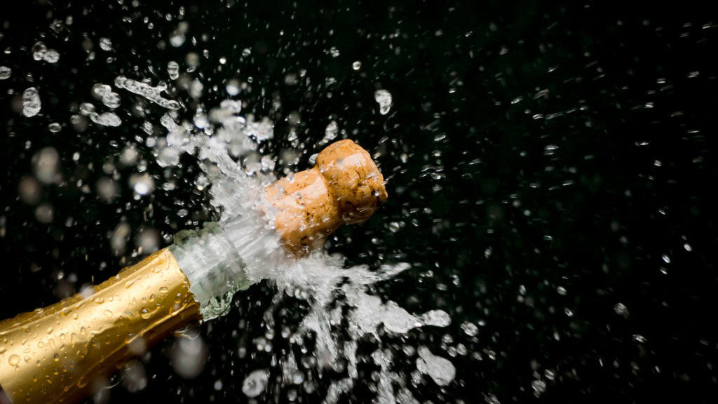 Das Bild zeigt einen Schaumwein, der gerade geöffnet wird. Der Korken ist bereits herausgesprungen und es spritzt überall. Die Flasche ist golden und mit Wassertropfen bedeckt. Der Hintergrund ist schwarz, was die Szene dramatisch hervorhebt. Die Aufnahme fängt den Moment der Feier und des Triumphes ein. Es wirkt dynamisch und lebhaft.