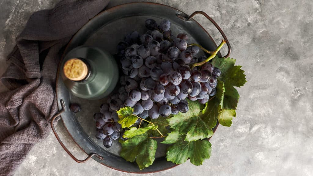 Traube der Syrah-Rebe mit grünen Blättern in einer runden Metallwanne neben einer grünen Weinflasche mit Korken auf grauem Untergrund