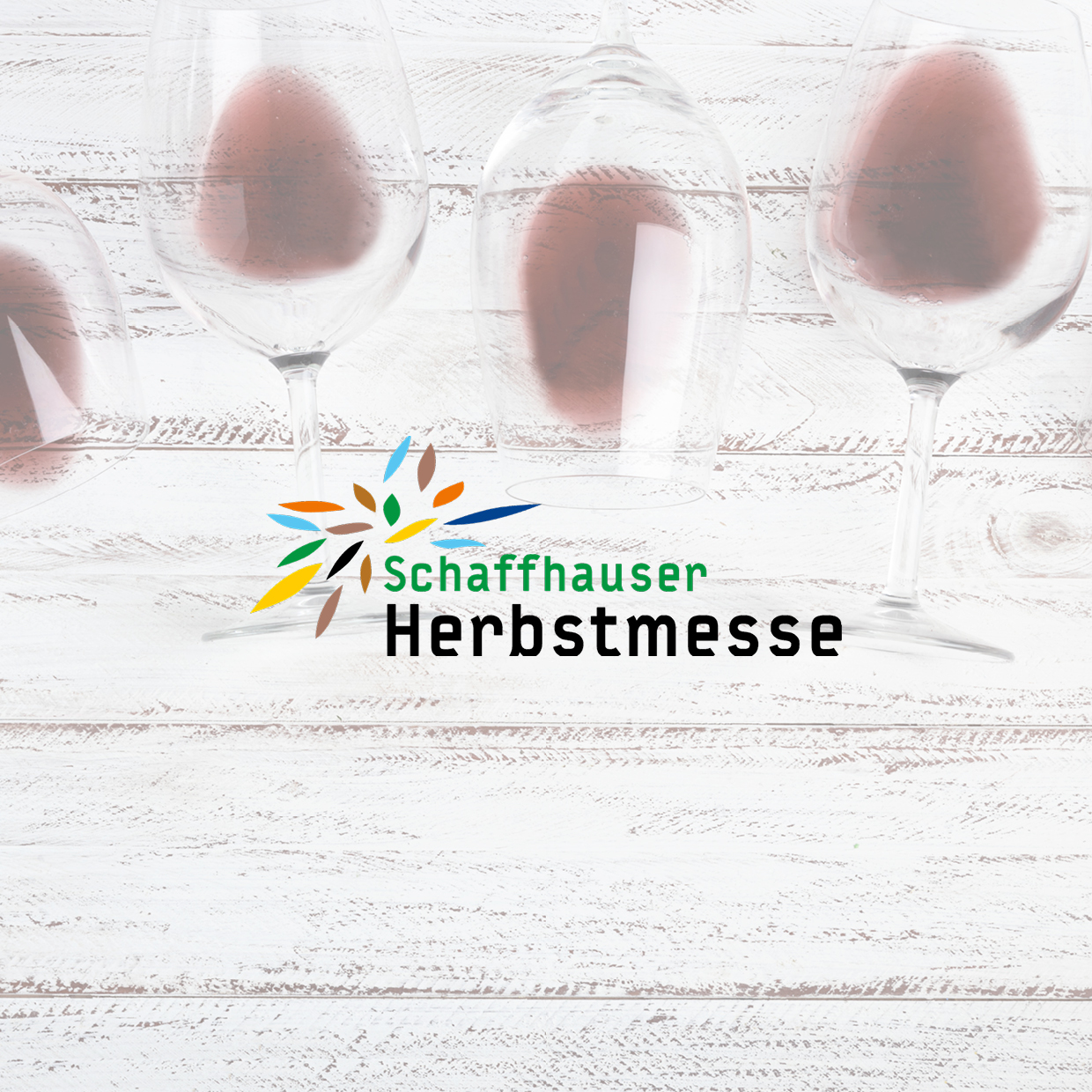 Mehrere Weingläser stehen auf einem hellen Holztisch mit dem Schriftzug 'Schaffhauser Herbstmesse' und einem bunten, stilisierten Blattlogo