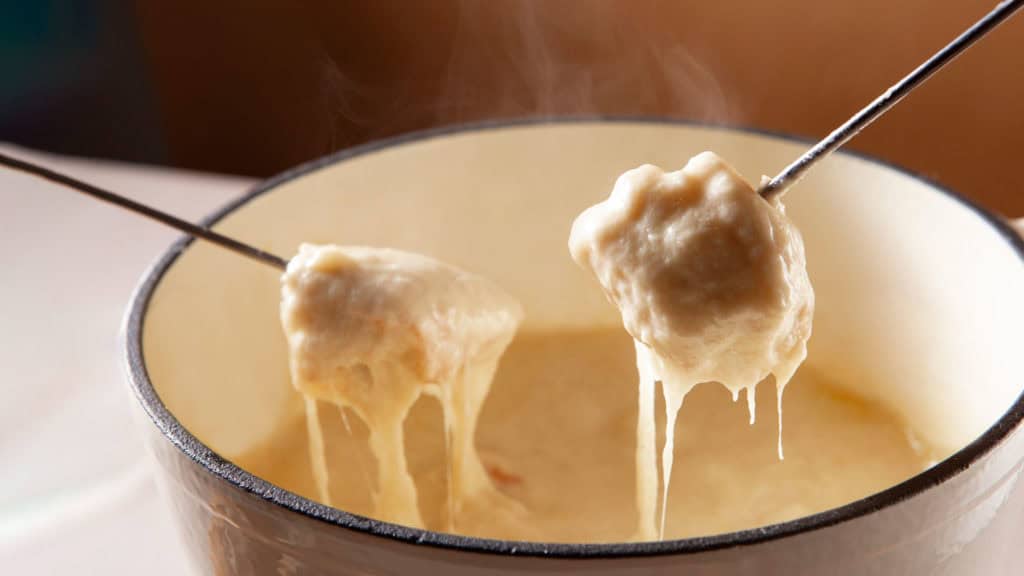 Käsefondue mit zwei Gabeln, Stück Brot in Käse getaucht, Wein im Hintergrund