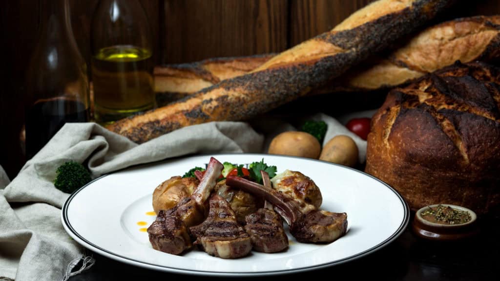 Gegrillte Lammkoteletts auf weissem Teller mit Brot, Pilzen und Olivenöl im Hintergrund