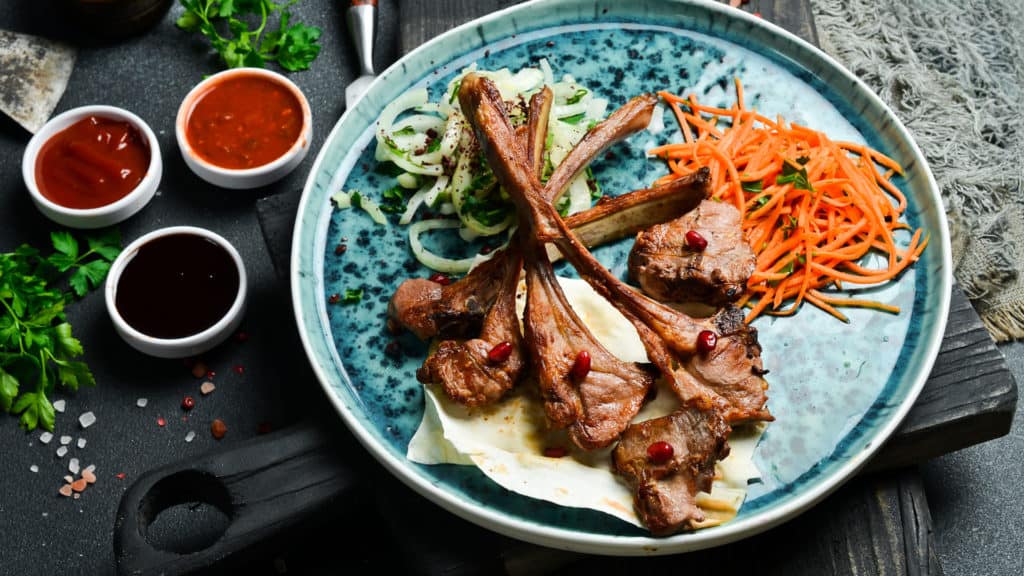 Gegrillte Lammkoteletts auf Fladenbrot mit Zwiebeln, geraspelten Karotten und drei kleinen Schälchen mit verschiedenen Sossen auf einem dunklen Holzbrett