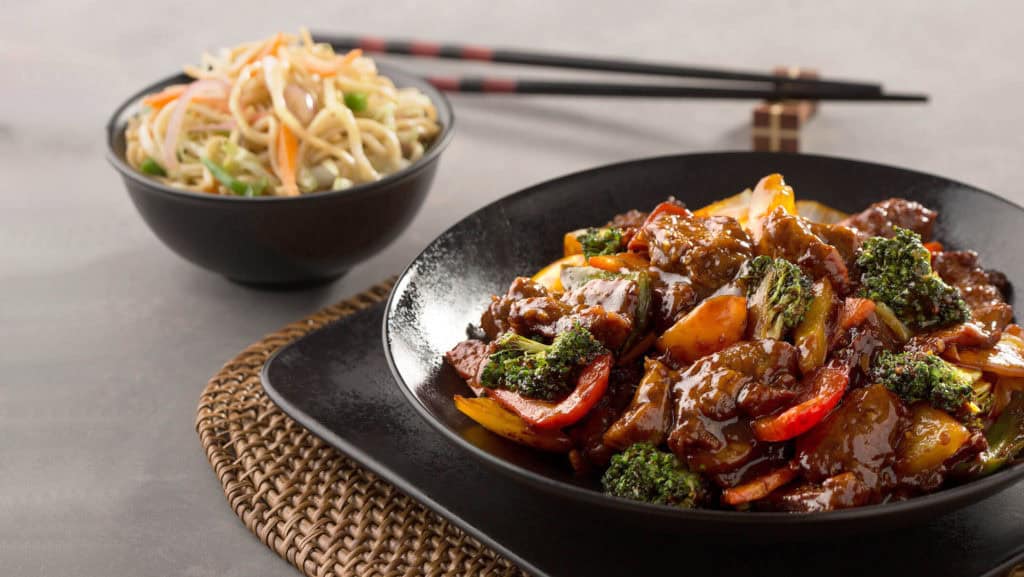 Schwarze Schale mit asiatisch gewürztem Lammfleisch und Gemüse neben einer Schale mit gebratenen Nudeln und Essstäbchen