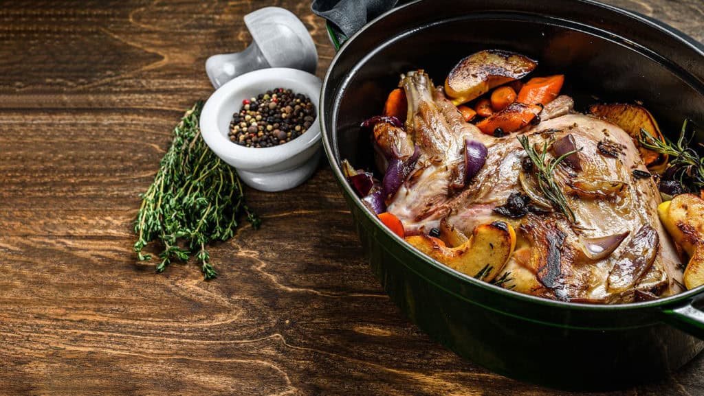 Geschmortes Lammfleisch mit Gemüse und Kräutern in einem schwarzen Topf auf Holztisch neben einem Mörser mit Pfefferkörnern und frischem Thymian