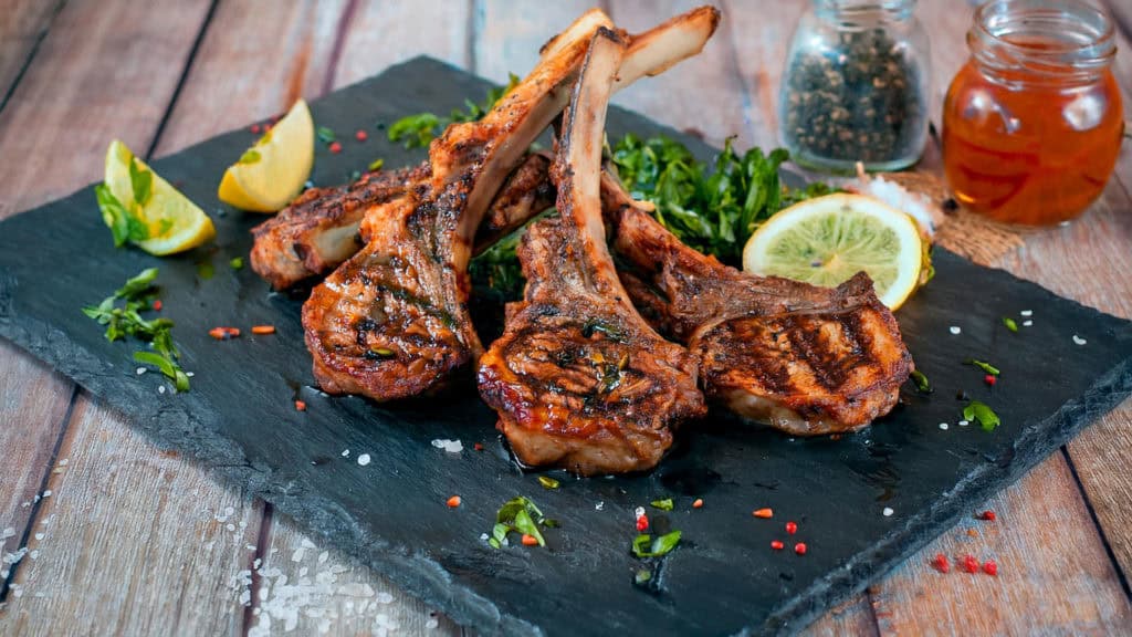 Mehrere gegrillte Lammkoteletts mit Kräutern auf schwarzer Steinplatte garniert mit Zitronenscheiben und Gewürzen auf Holztisch