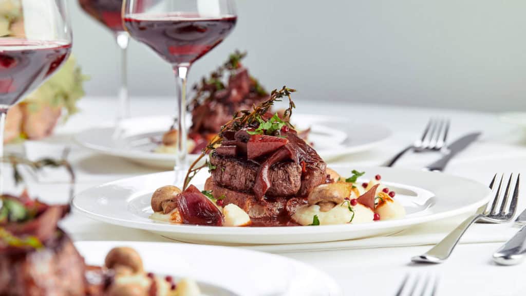 Wein & Wild: Rotwein im Weinglas neben Wildgericht auf Holztisch