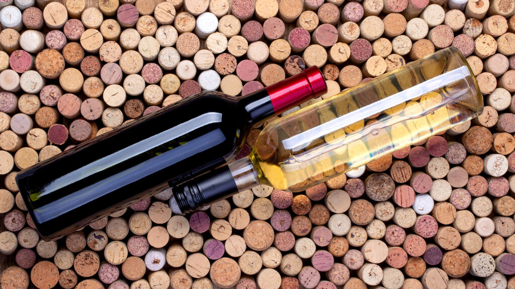 Zwei Weinflaschen, eine mit rotem und eine mit weissem Wein, liegen nebeneinander auf einer Oberfläche aus vielen Weinkorken. Fokus auf Standardweinflasche 75cl Flaschengrösse