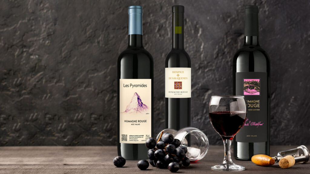 Drei Flaschen Rotwein: Les Pyramides Humagne Rouge, Hospices de Salquenen Humagne Rouge und Humagne Rouge, neben einem Glas Rotwein und Trauben.