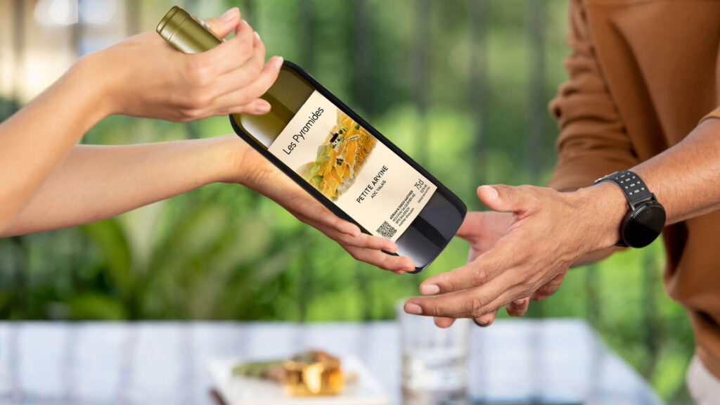 Zwei Hände übergeben eine Weinflasche mit Etikett 'Les Pyramides Petite Arvine' vor unscharfem Hintergrund mit Glas und Teller