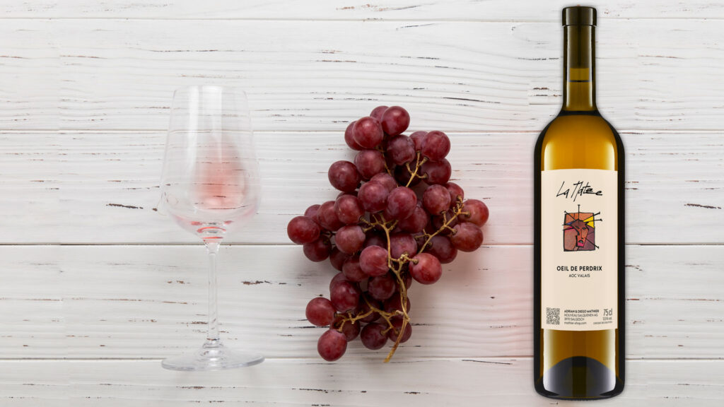 Rosé wine Oeil-de-Perdrix La Matze with red grapes and a wine glass on a wooden surface.