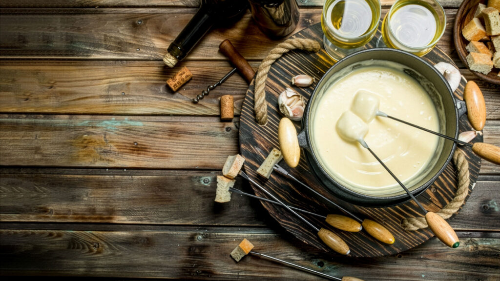 Cheese fondue 5 Cheese fondue in glass with wooden handle, two forks with pieces of bread, two white wine glasses, wine bottle, cork and garlic on wooden board and wooden table.