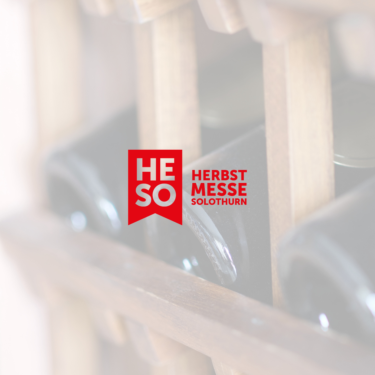 Rotes Logo mit den Buchstaben HESO und dem Text HERBST MESSE SOLOTHURN vor unscharfem Hintergrund mit Weinflaschen und Holzlatten