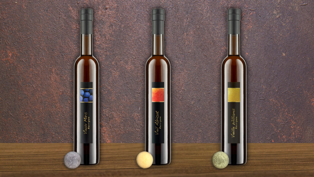 Drei schlanke Flaschen mit dunklem Inhalt und farbigen Etiketten stehen auf einem Holztisch mit Pralinen vor einer braunen Wand.