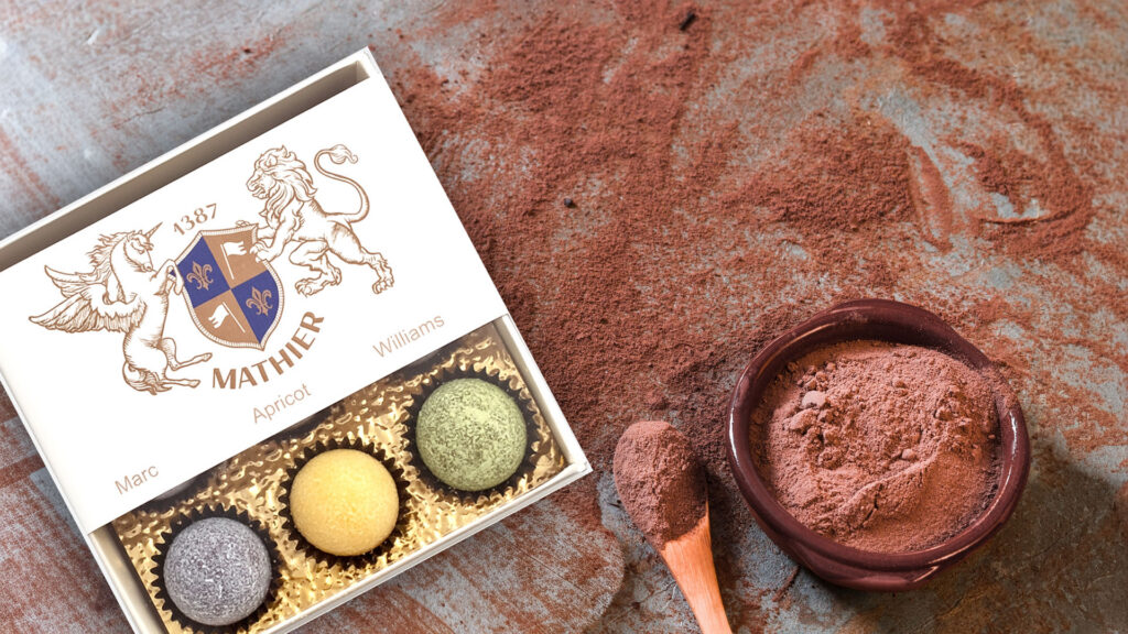 Pralinenverpackung mit drei Pralinen in verschiedenen Farben neben einem kleinen Tontopf mit Kakaopulver und einem Löffel mit Kakaopulver.