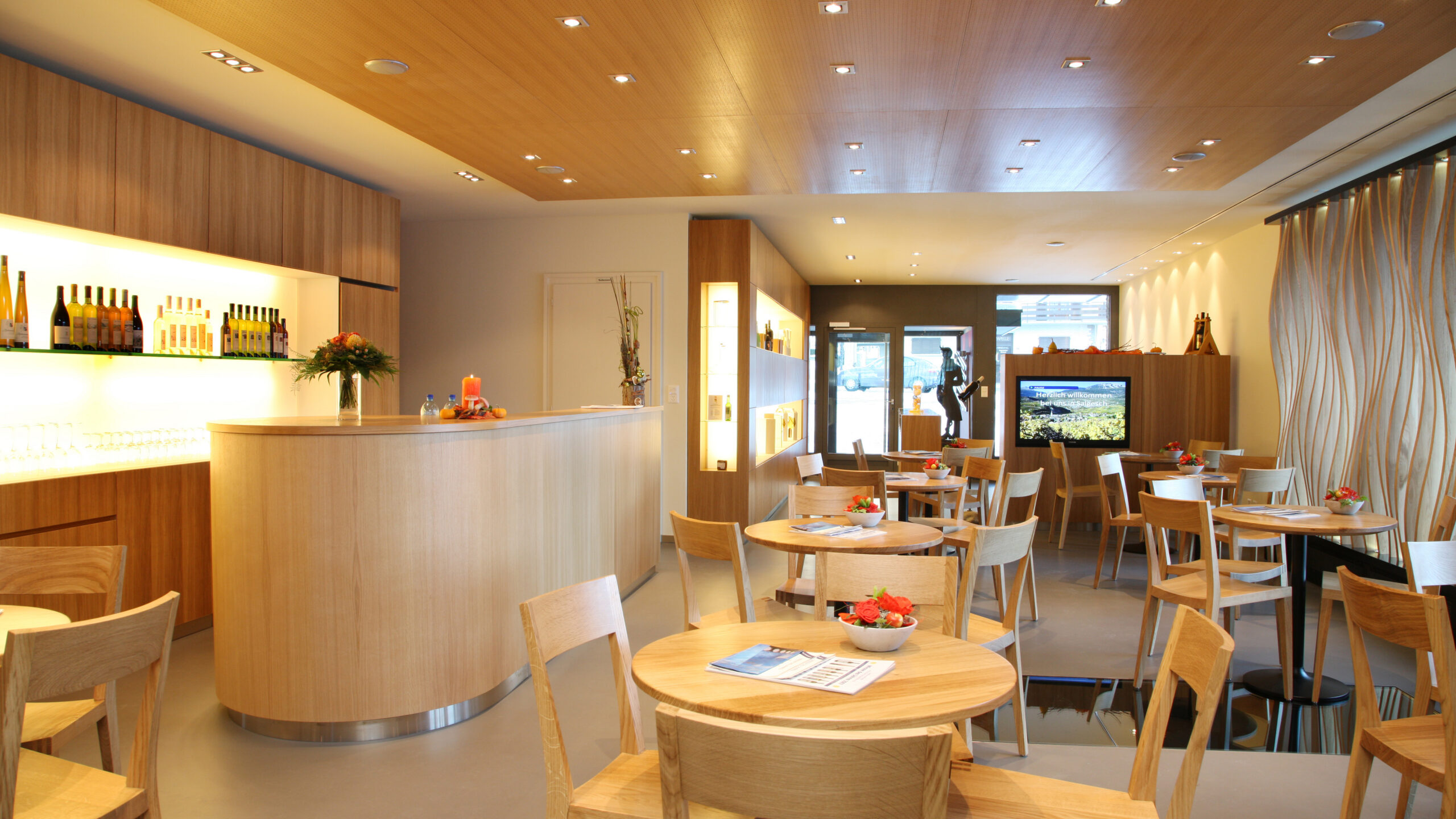 Modern guest room in the Adrian & Diego Mathier winery with light-colored wooden tables and chairs, bar with illuminated shelves and decorative flower arrangements