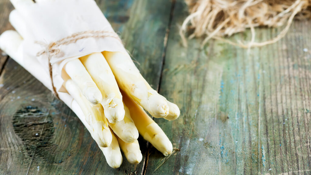 Thema Wein zu Spargel: Ein Bündel weisser Spargelstangen, locker in ein helles Tuch gewickelt und mit einer Schnur gebunden, liegt auf einer verwitterten Holzoberfläche