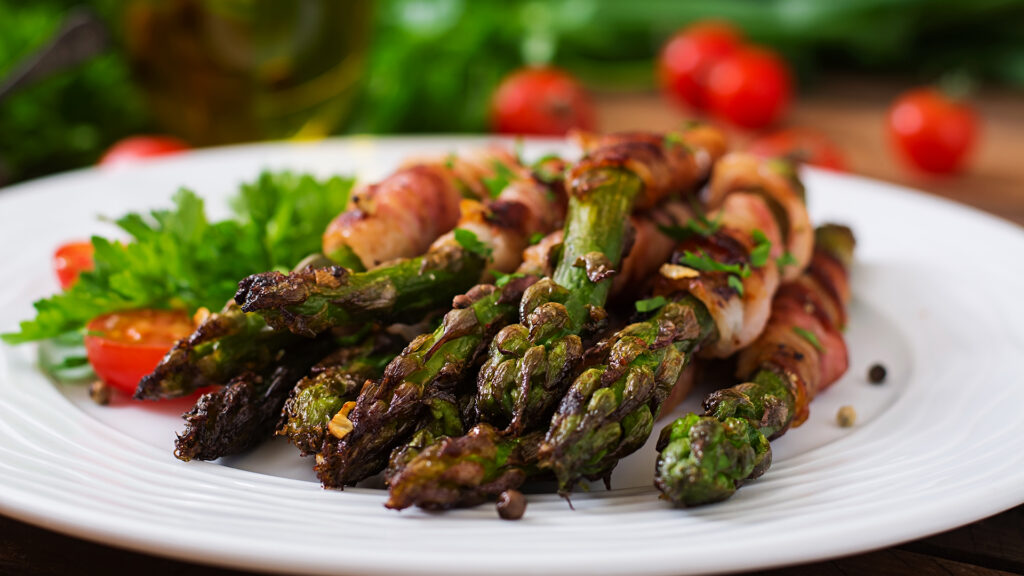 Thema Weine zu Spargel: Grüne Spargelstangen, die mit Schinken umwickelt und auf einem weissen Teller mit Petersilie und halbierten Kirschtomaten angerichtet sind