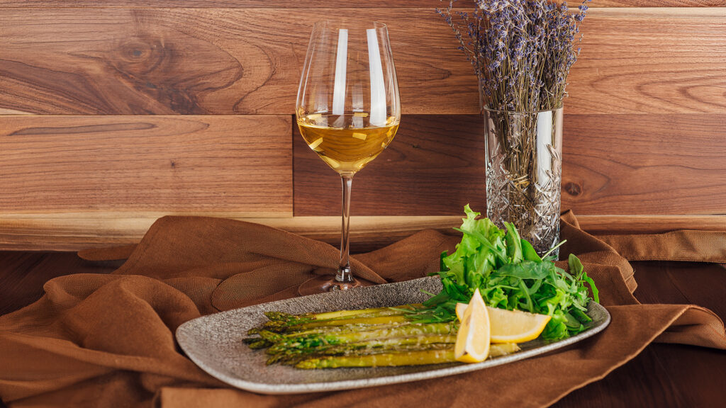 Grüner Spargel mit Zitronenspalten und Salat auf einem grauen Teller neben einem Glas Weisswein und einem Glas mit getrockneten Blumen vor einer Holzverkleidung
