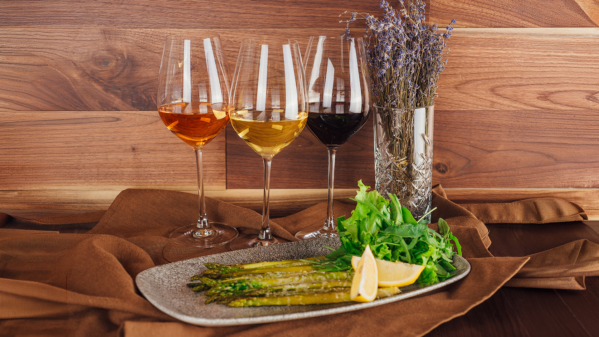 Thema Wein zu Spargel: Drei Weingläser mit unterschiedlichem Wein, Teller mit grünem Spargel, Zitronenspalten und Salat, daneben Vase mit getrockneten Blumen auf braunem Tuch vor Holzwand