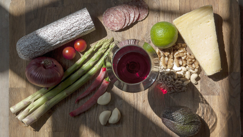 Grüner Spargel, rote Chilischoten, rote Zwiebel, Kirschtomaten, Knoblauchzehen, Scheiben Salami, Limette, Käse, Nüsse, Avocado und ein Glas Rotwein auf Holzbrett