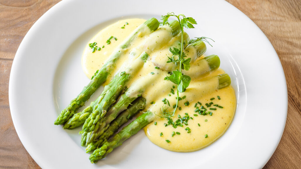 Grüner Spargel mit Sauce Hollandaise und Kräutern auf weissem Teller auf Holztisch