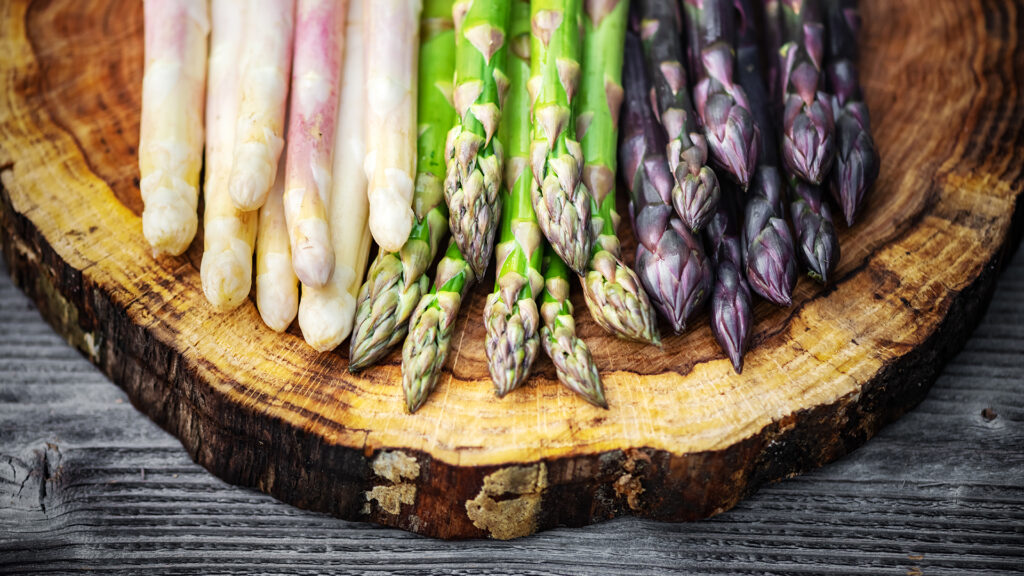 Thema Wein und Spargel: Drei Sorten Spargel in weiss, grün und violett liegen nebeneinander auf einer runden Holzscheibe.