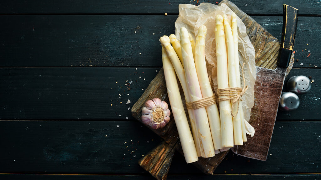 Thema Wein zu Spargel: Zwei Bündel weißer Spargel auf einem Holzbrett mit einem Messer, Knoblauchknolle und Salz- und Pfefferstreuer auf dunklem Untergrund